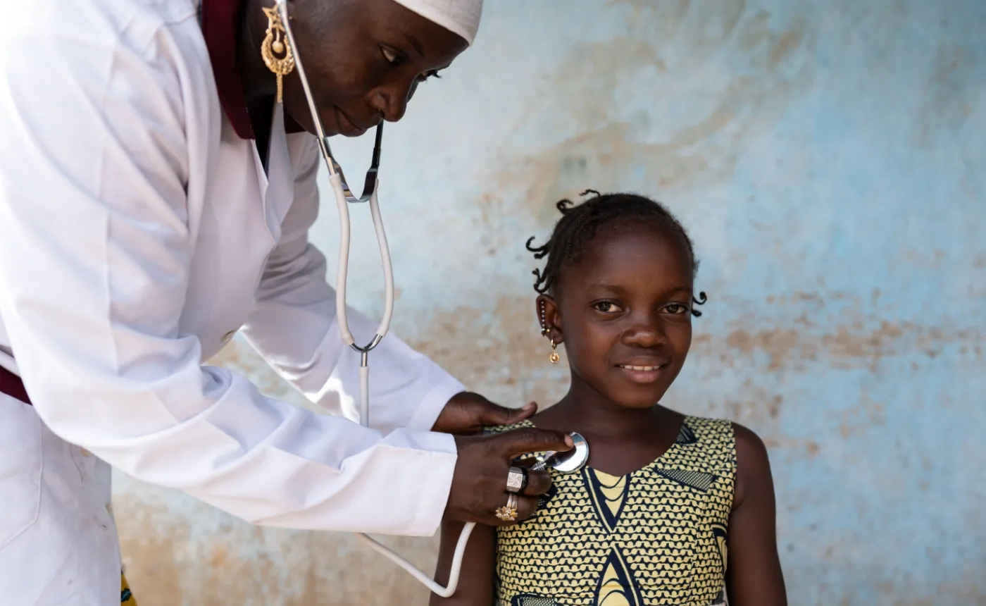 Ein Foto zeigt eine lächelnde junge Ärztin, erkennbar an ihrem weissen Laborkittel und ihrer weissen Kappe, die ein Stethoskop verwendet, um das Herz eines lächelnden Mädchens abzuhören. Das Mädchen trägt ein traditionelles, gemustertes Kleid und Ohrringe. Sie stehen beide vor einer Wand mit abblätternder blauer Farbe, was einen einfachen medizinischen Kontext vermuten lässt.